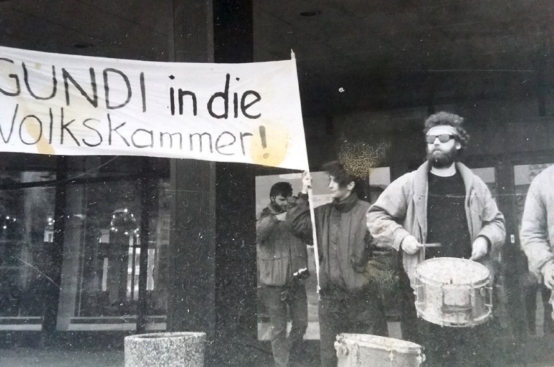 Black and white image of a protest. Bearded man in foreground beating a drum, others behind holding banner reading 'GUNDI in die Volkskammer!'